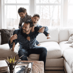 Man and his two sons playing on a white couch in a stylish living room. Family having fun in new home at Rosewood at Secord in Edmonton