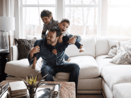 Man and his two sons playing on a white couch in a stylish living room. Family having fun in new home at Rosewood at Secord in Edmonton